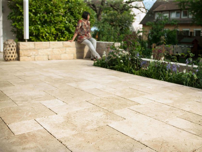 Frau sitzt auf Natursteinmauer, davor eine Terrasse mit hellen Travertinplatten und ein Blumenbeet