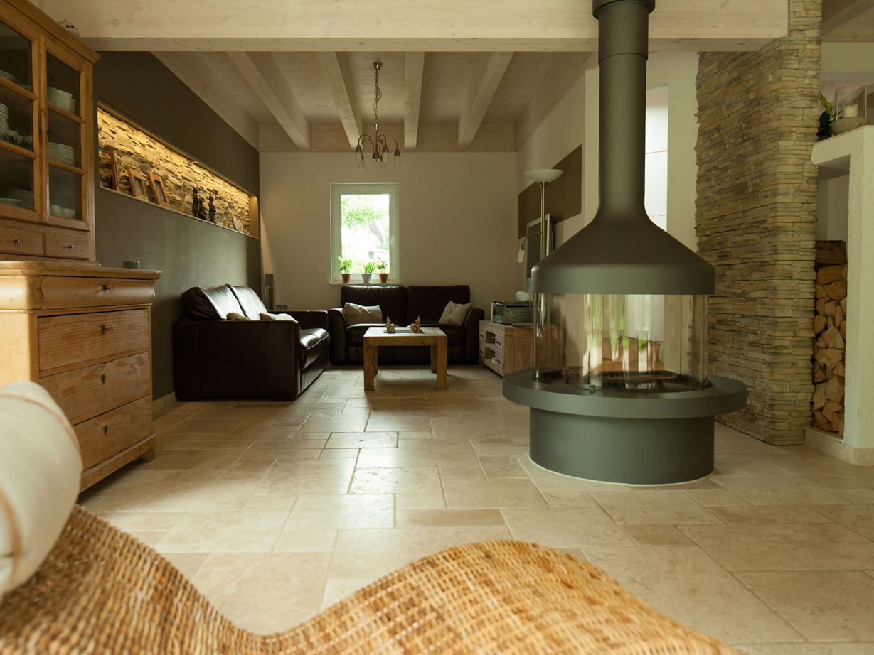 Wohnzimmer mit alleinstehendem Kamin und Sitzecke auf Travertinfliese Light, im Vordergrund ein Korbsessel