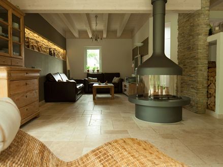 Wohnzimmer mit alleinstehendem Kamin und Sitzecke auf Travertinfliese Light, im Vordergrund ein Korbsessel