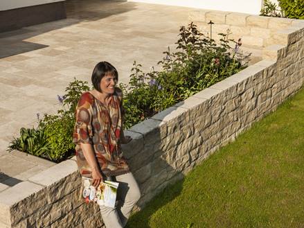Frau lehnt an einer Natursteinmauer, im Hintergrund eine Terrasse mit Travertinplatten, vorne eine Rasenfläche