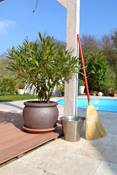 Ein Besen lehnt an einem Pergolapfosten auf einer Natursteinterrasse, im Hintergrund ein Pool und links eine Topfpflanze